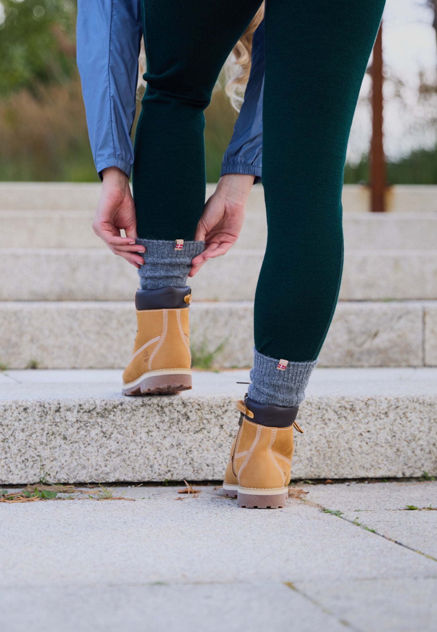 Person adjusts gray alpaca wool socks over green leggings with tan boots on stone steps.