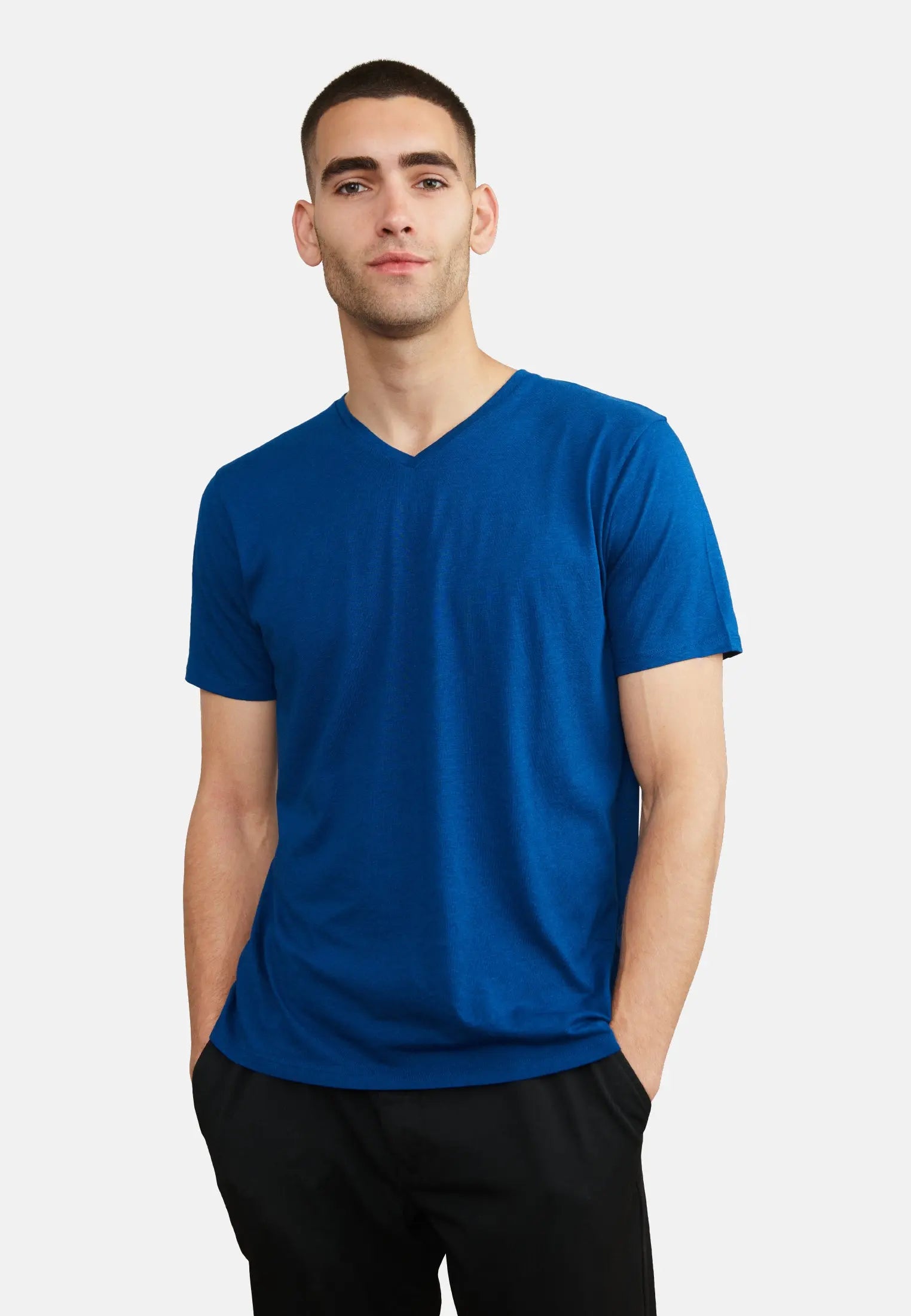 Man wearing a blue V-neck T-shirt with hands in pockets, standing against a white background.