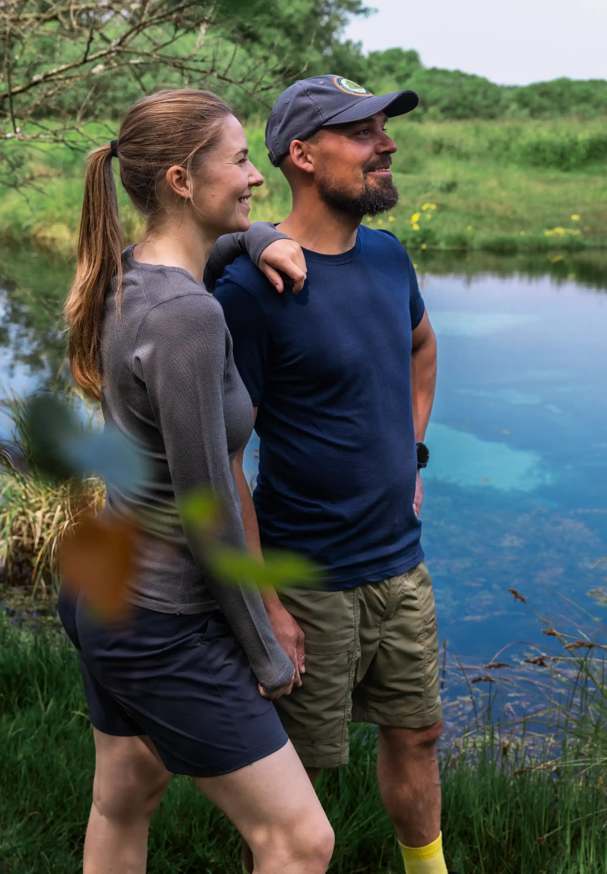 Man in blue T-shirt and cap stands by a pond with a woman, surrounded by green foliage.