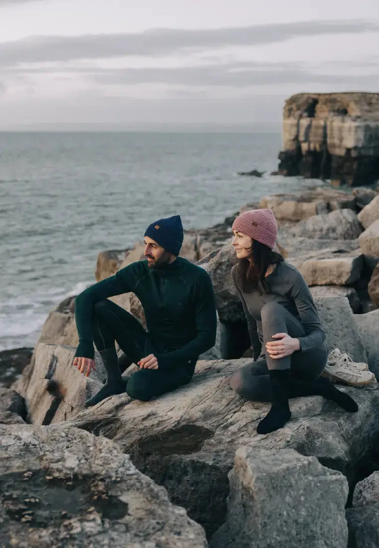 Two people in merino wool base layer pants on rocky shore, wearing beanies with ocean in background.