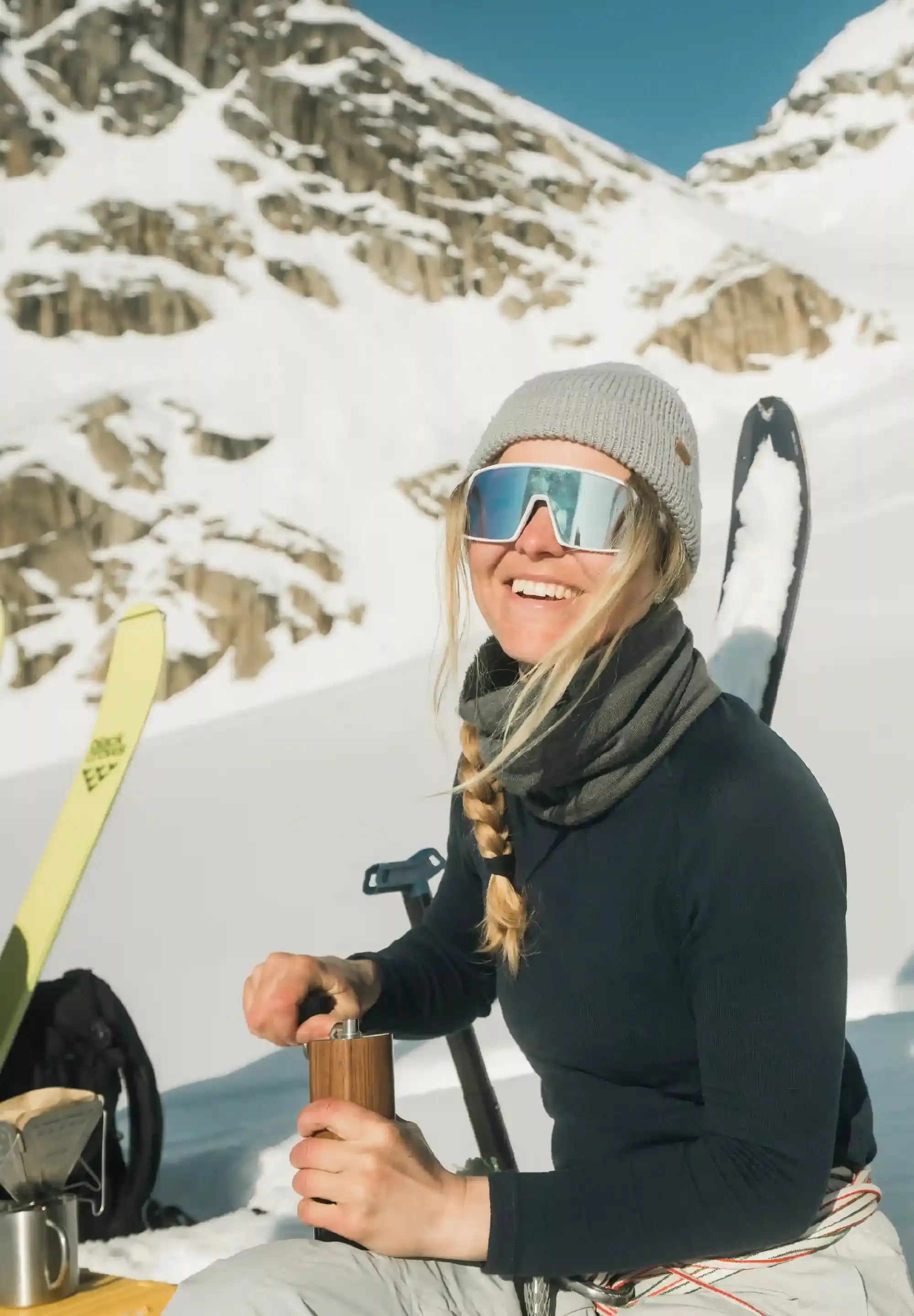 Woman in merino base layer, beanie, and sunglasses, smiling in snowy mountains with skis.