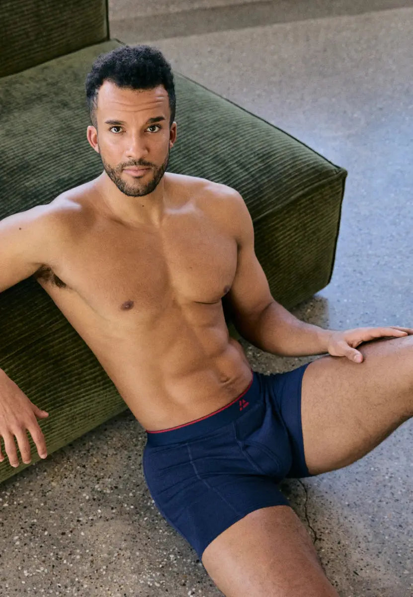 Man wearing navy bamboo trunks with red waistband, sitting on the floor beside a green sofa.