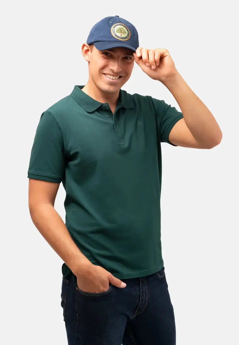 Person in a blue cap with emblem, green polo shirt, and jeans, smiling and holding cap brim.