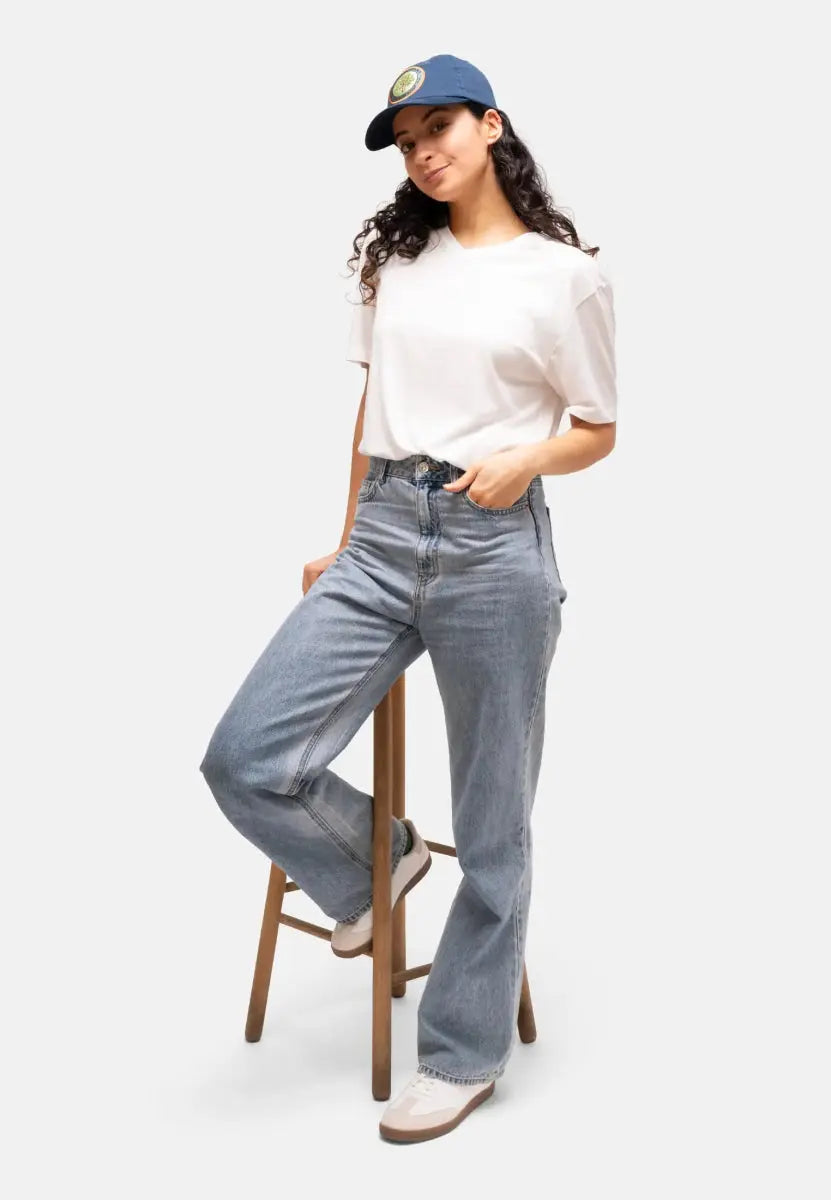 Person in navy cap, white t-shirt, light jeans, leaning on a wooden stool.