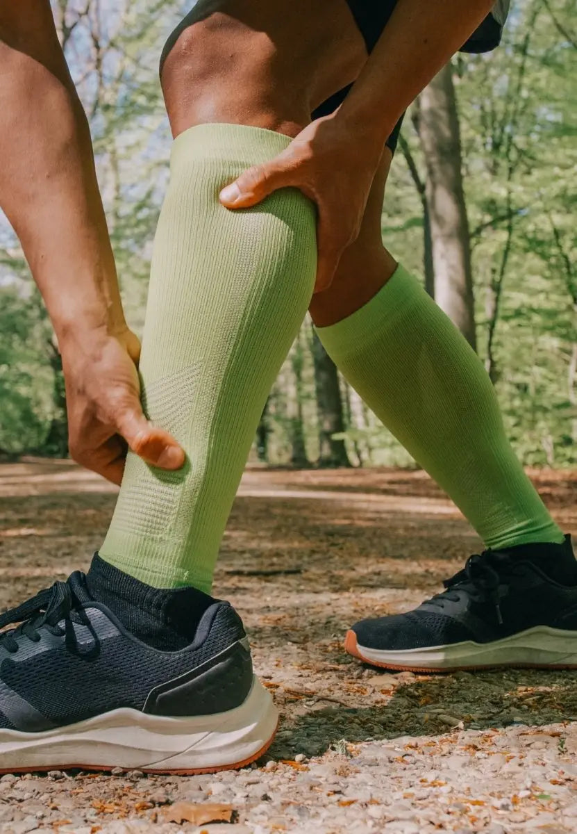 Person outdoors adjusting green calf compression sleeves, wearing black sneakers on a forest path.