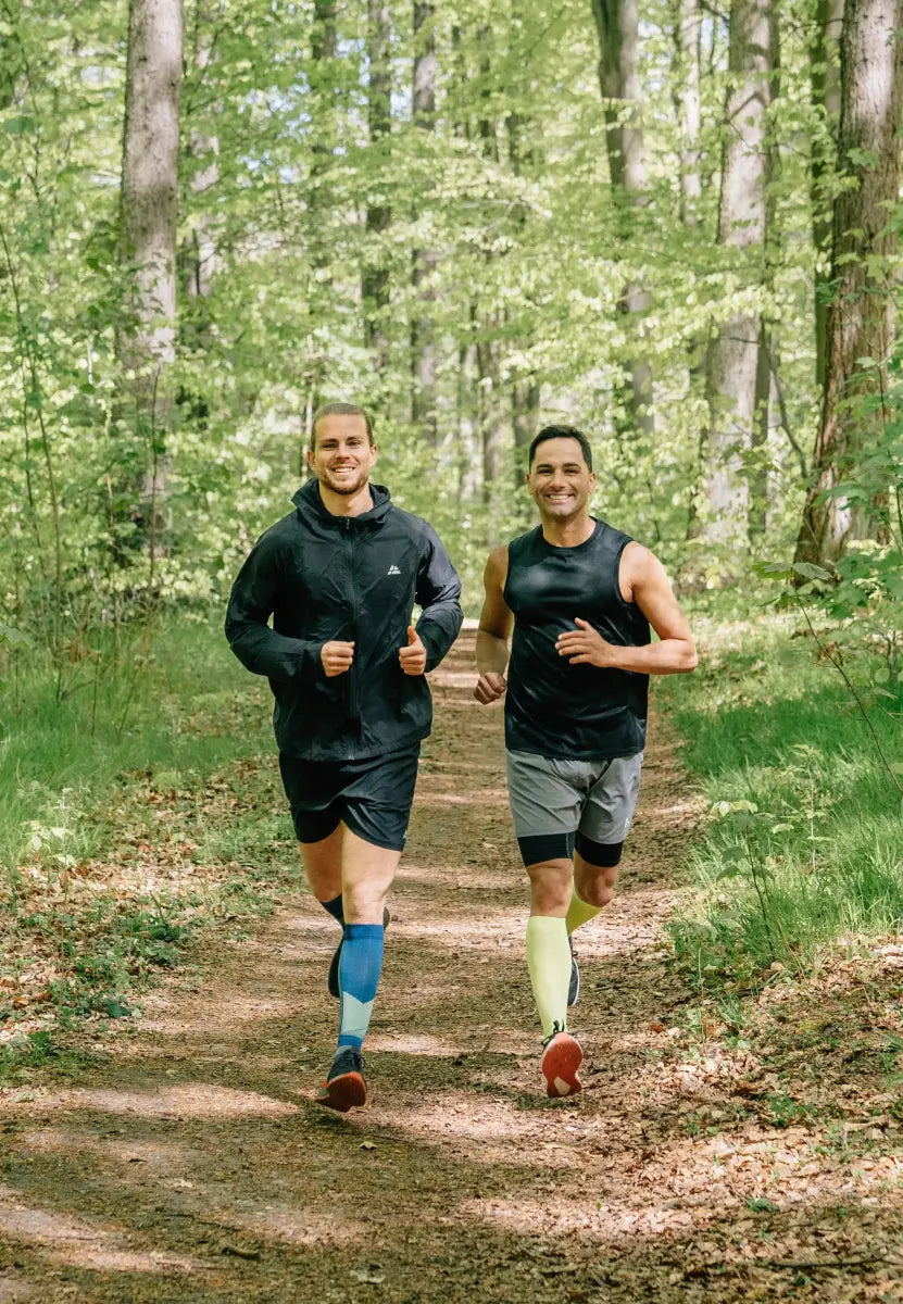 Two men jogging on a forest path, wearing blue and yellow calf compression sleeves.