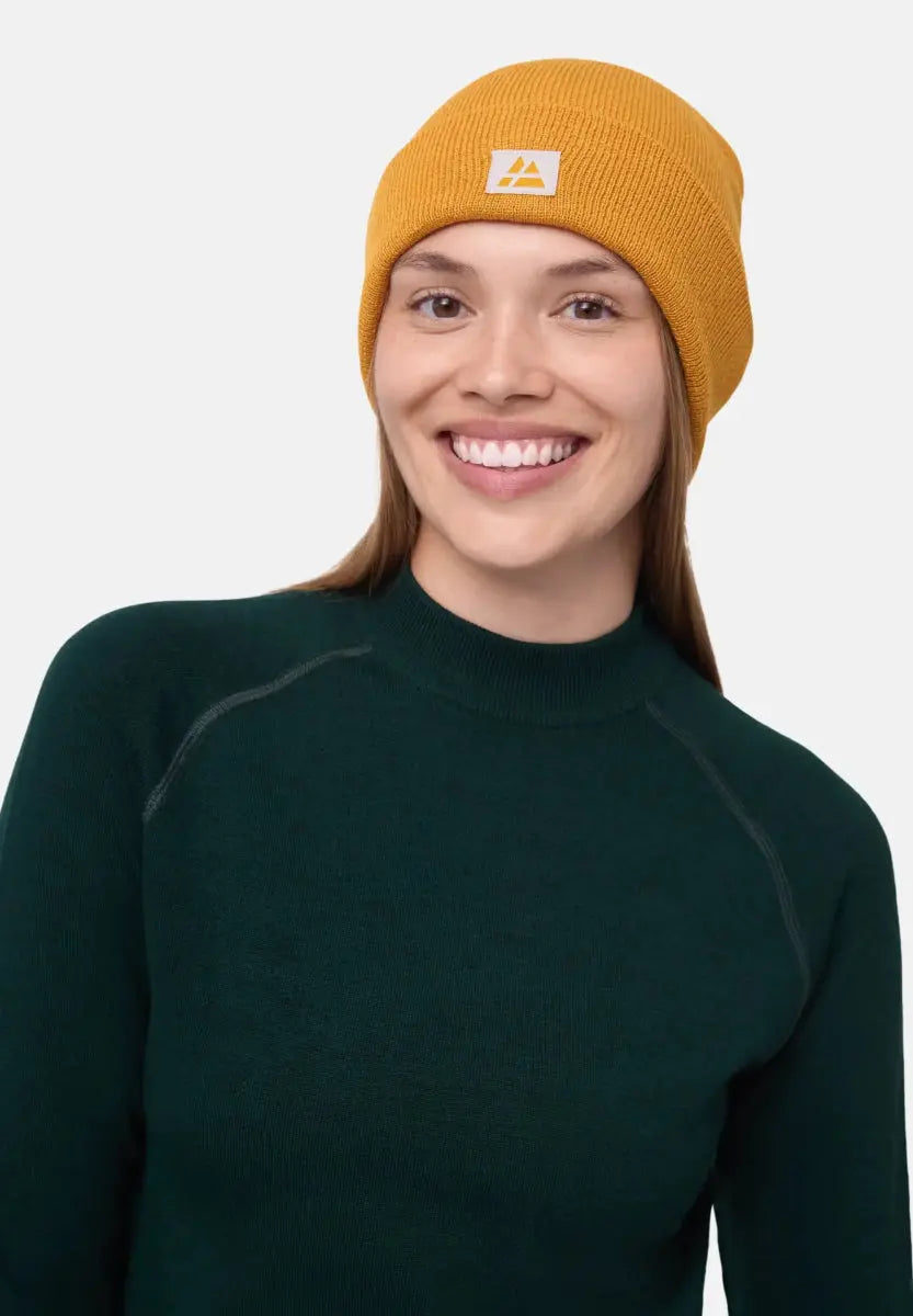 Person wearing a mustard yellow beanie with logo, smiling, in a dark green top.