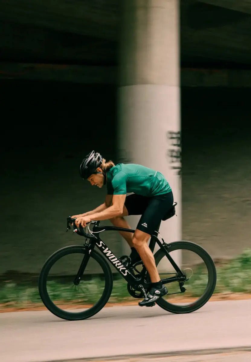 Cyclist in green jersey and black bib shorts riding a black bike under a bridge.