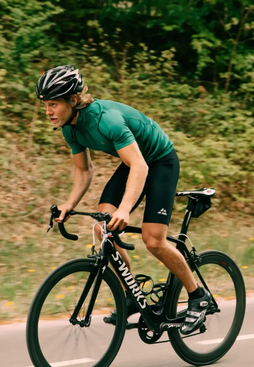 Cyclist in green jersey and black bib shorts, riding on a road with greenery in the background.