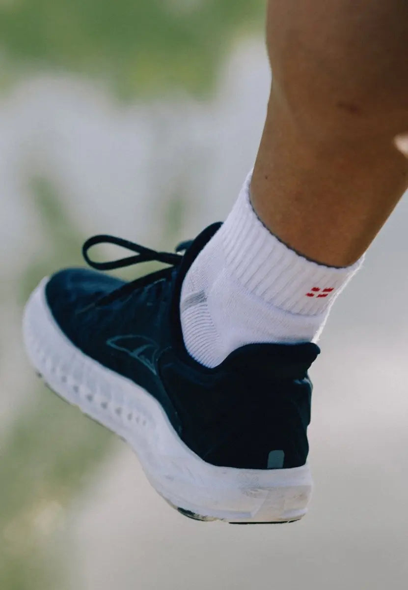 Foot in white cotton quarter sock with red logo, wearing black athletic shoe, blurred background.