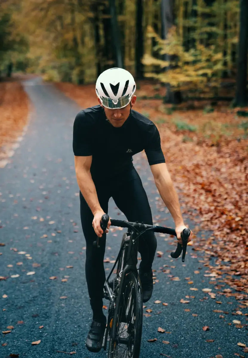 Man cycling on a forest road, wearing a white helmet and black bib pants, surrounded by autumn leaves.