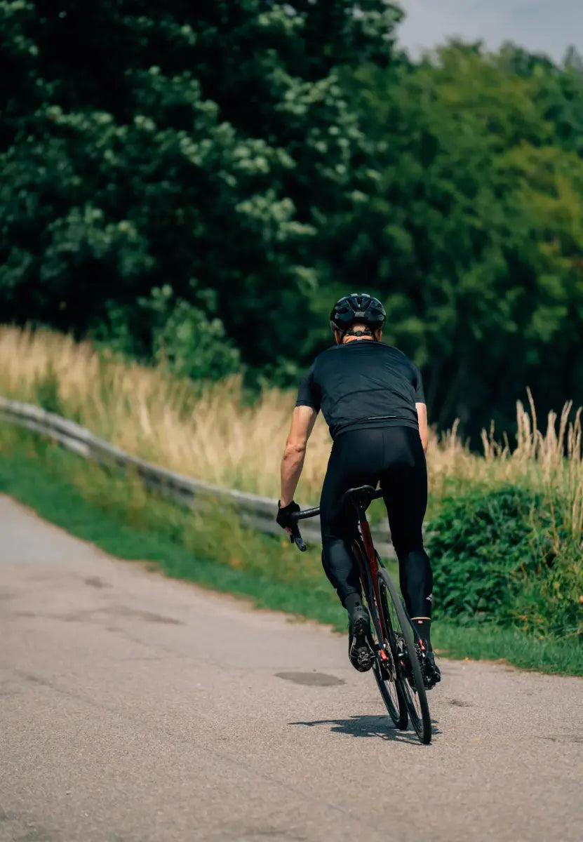 Cyclist in black bib pants and helmet riding on a road with lush green surroundings.