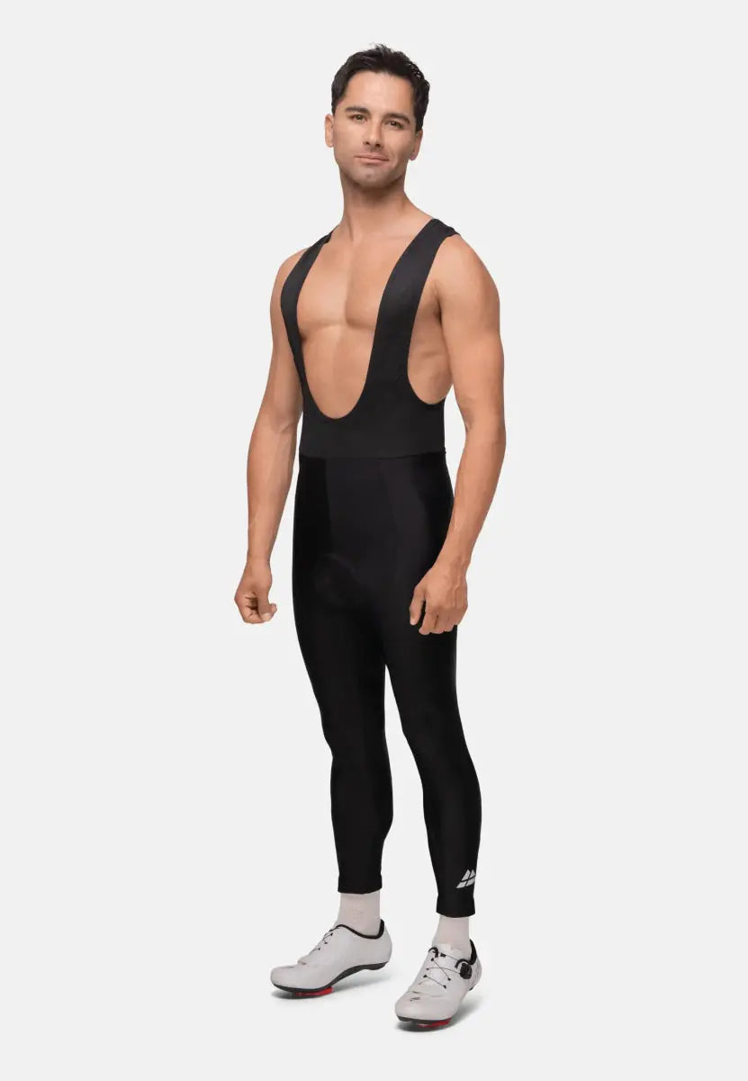 Man wearing black cycling bib pants, white shoes, and socks on a plain background.
