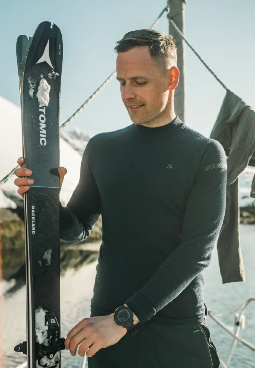Man in black merino wool base layer holding skis with snowy mountains in the background.