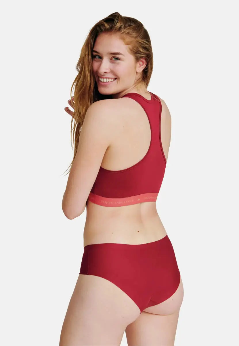 Person in red sports bra and hipster panties, smiling over shoulder against a white background.