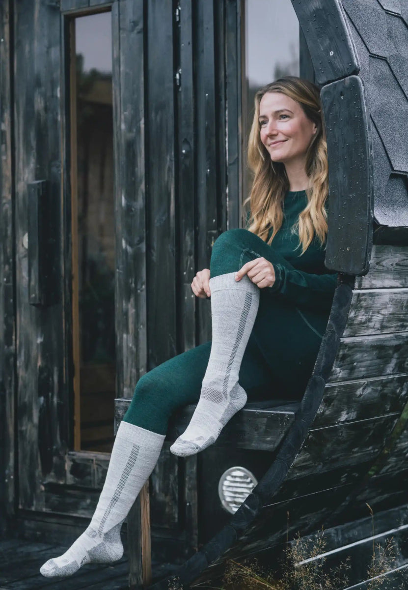 Person wearing knee-high merino hiking socks, sitting outdoors on a wooden structure, dressed warmly.