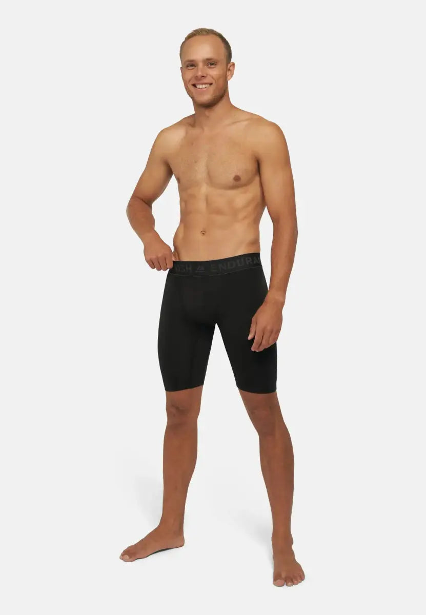 Shirtless man smiling, wearing black compression shorts, standing with a hand on his hip.
