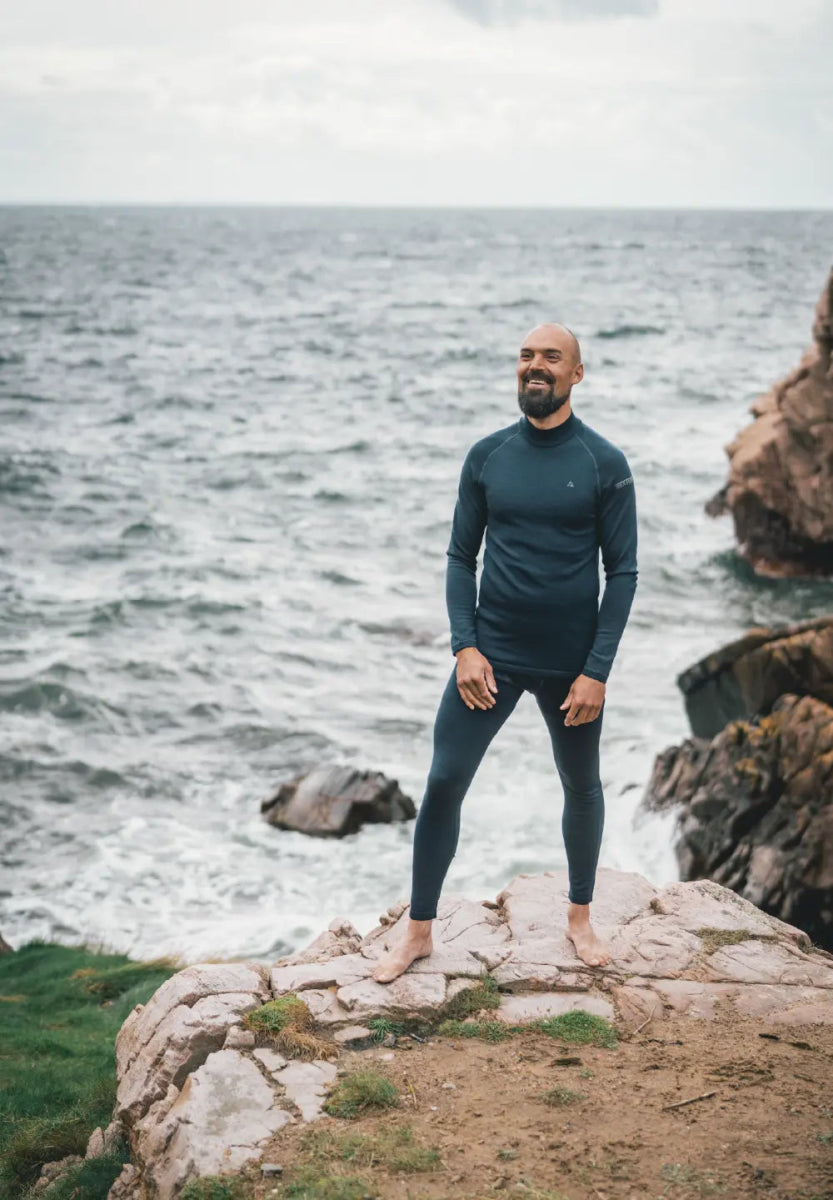 Man in dark merino base layer tights stands on a rocky cliff by the ocean, smiling.