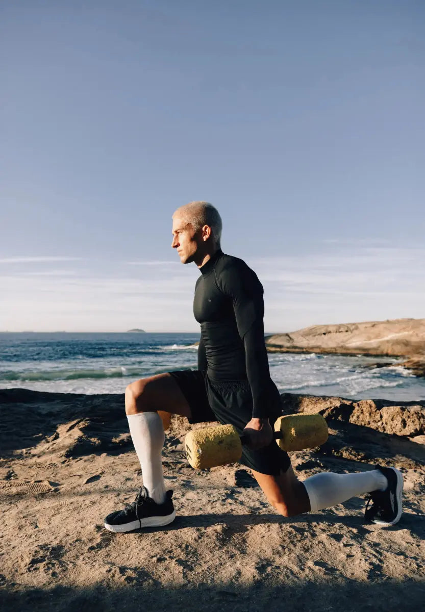 Man in black compression shirt lunging with a yellow dumbbell on rocky beach, ocean in background.