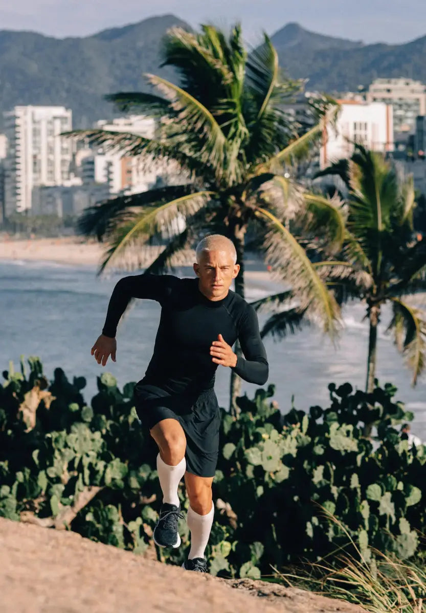Man running uphill in black compression shirt near beach with palm trees and mountains in background.