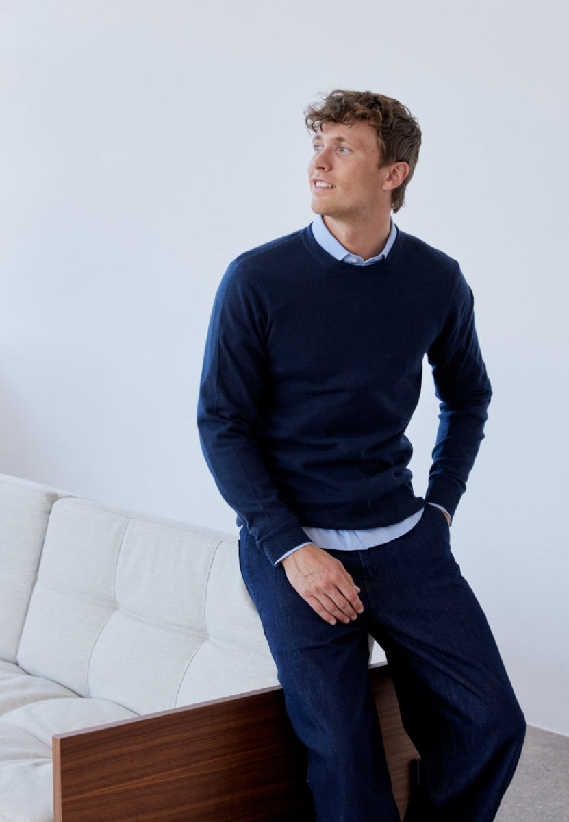 Man in navy sweater over light blue shirt, sitting on couch, looking to the side.