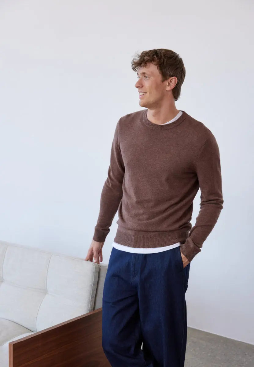 Man wearing a brown sweater and blue pants, standing in a light, minimalistic room.