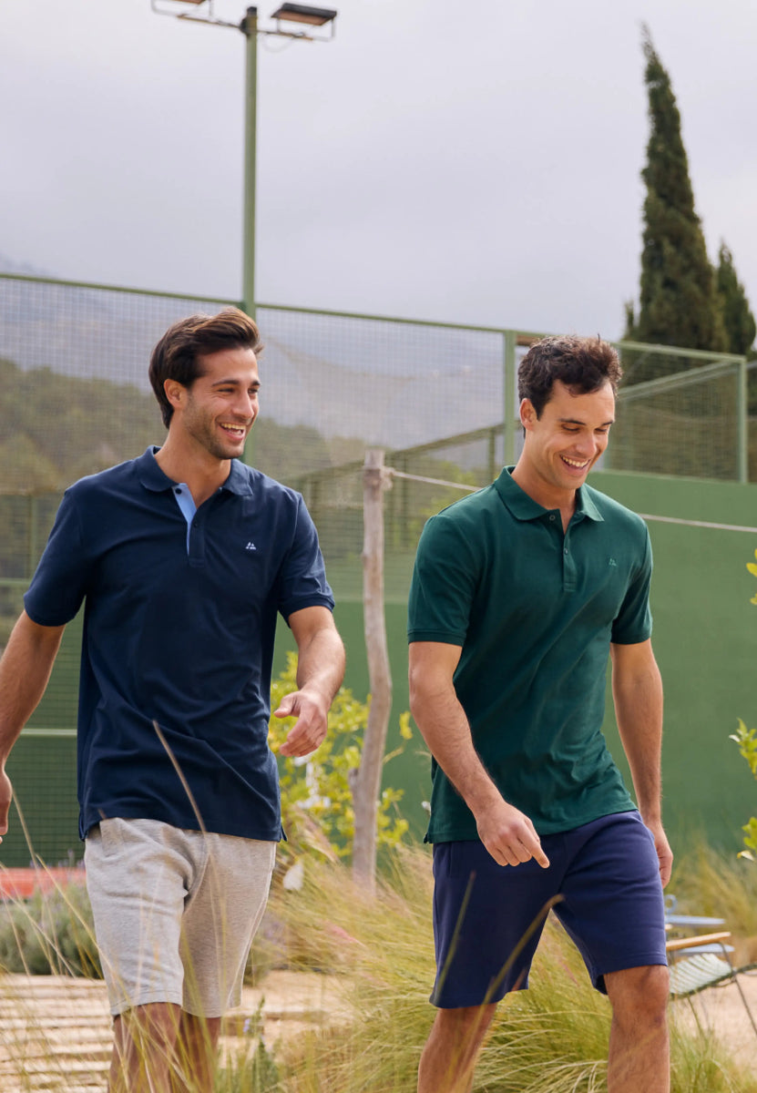 Two men walking outdoors, wearing navy and green organic cotton polo shirts, smiling.