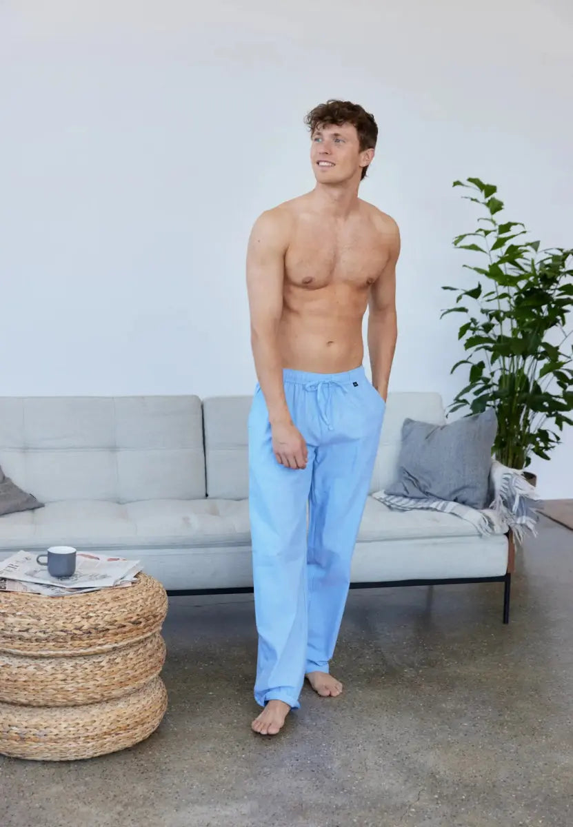Shirtless man in light blue pyjama pants, standing in a stylish living room with plants and a sofa.