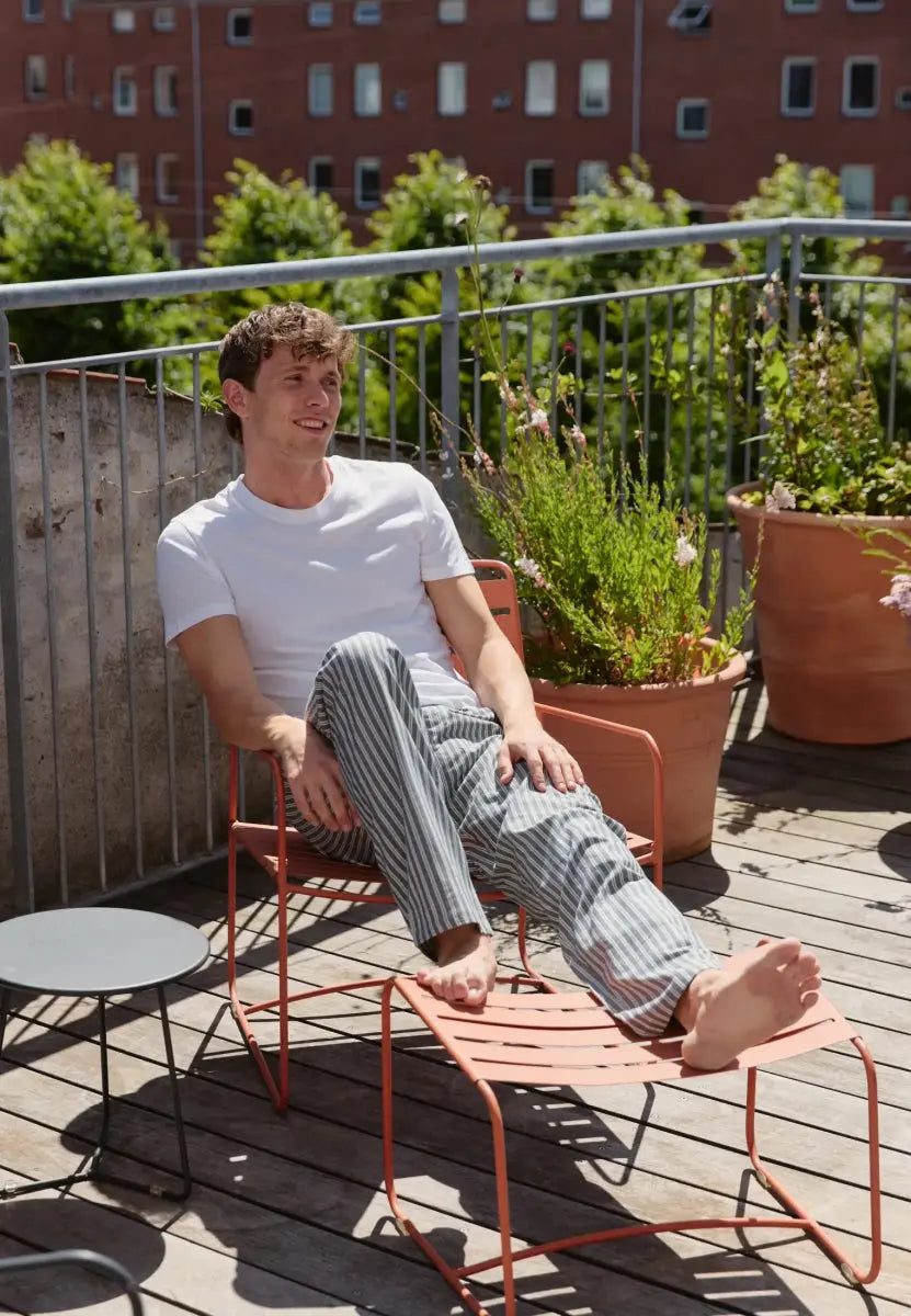 Man in striped pyjama pants and white T-shirt relaxing on a terrace with plants and metal furniture.