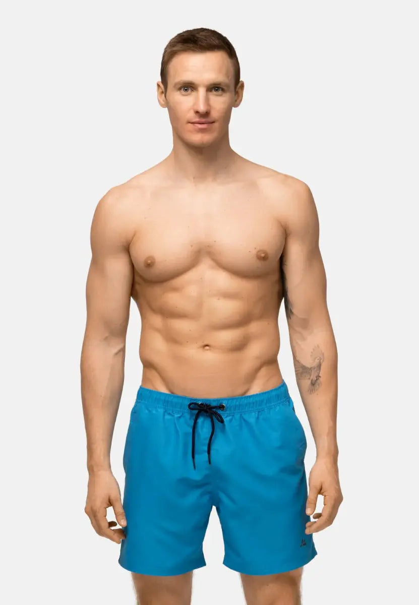 Shirtless man wearing blue quick-dry swim trunks with black drawstring and tattoo on left arm.