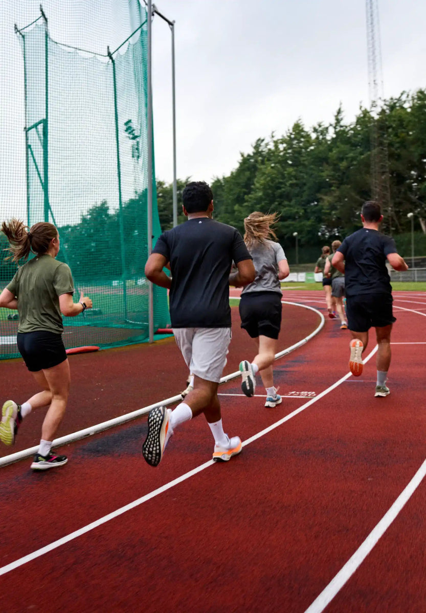 People running on a red track wearing athletic clothing, with trees and a netted area in the background.