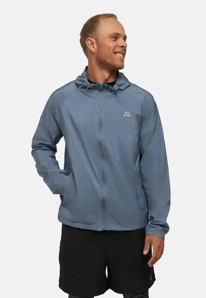 Man in a light blue hooded windbreaker jacket and black shorts, smiling with hands in pockets.