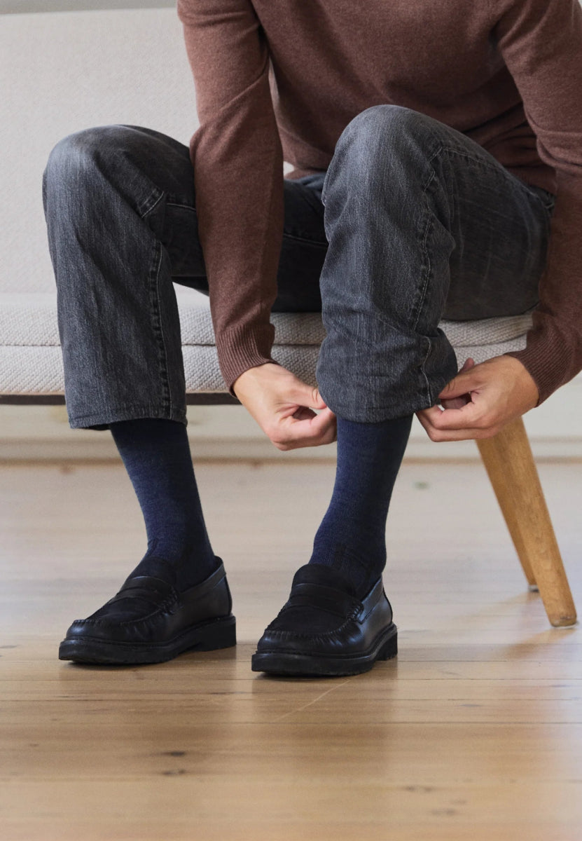 Person in jeans adjusts dark blue socks with black shoes, seated on a light-colored bench.