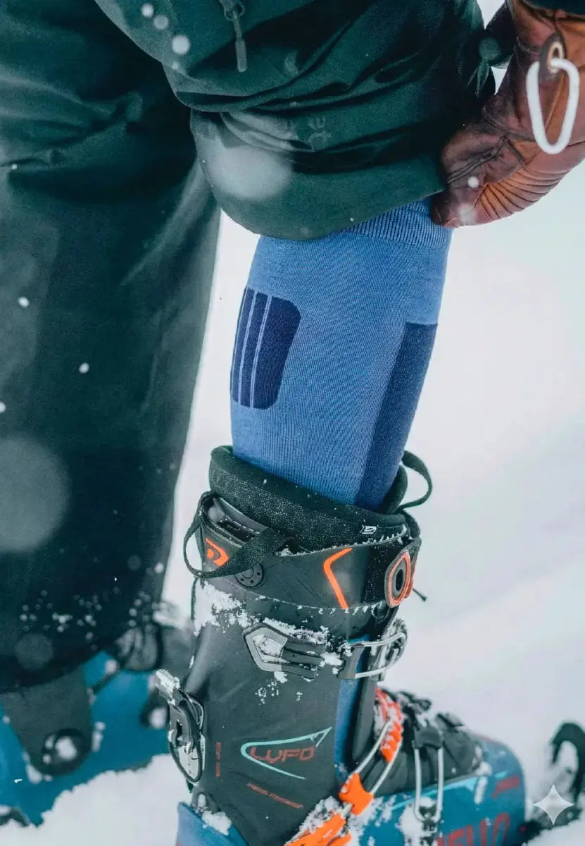 Blue Merino ski sock being adjusted inside a black ski boot with snow, showing winter gear.