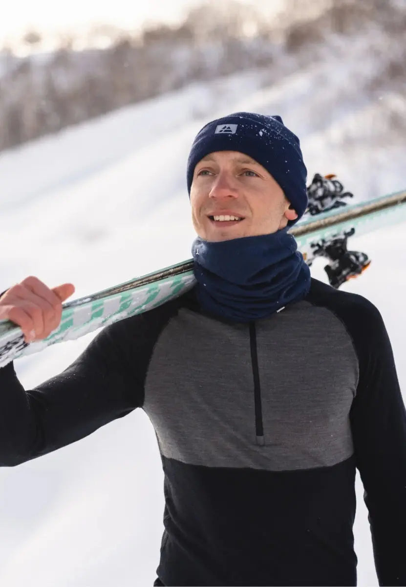 Man in black and gray merino wool shirt, carrying skis, wearing a navy beanie and neck gaiter in snow.