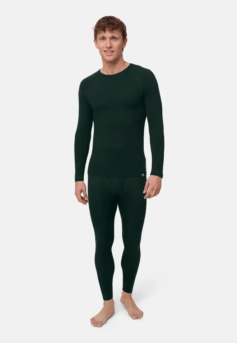 Man in dark green merino wool base layer set with long sleeves and leggings, standing barefoot.