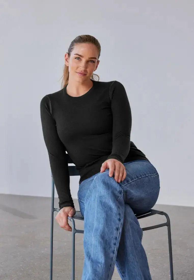 Woman sitting, wearing a black merino wool base layer top and blue jeans, posing casually on a chair.