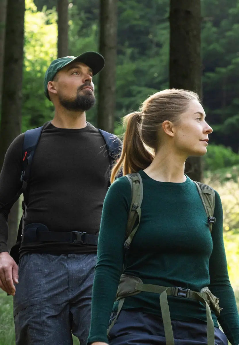 Two hikers in forest wearing merino wool base layer shirts, one black, one green, with backpacks.