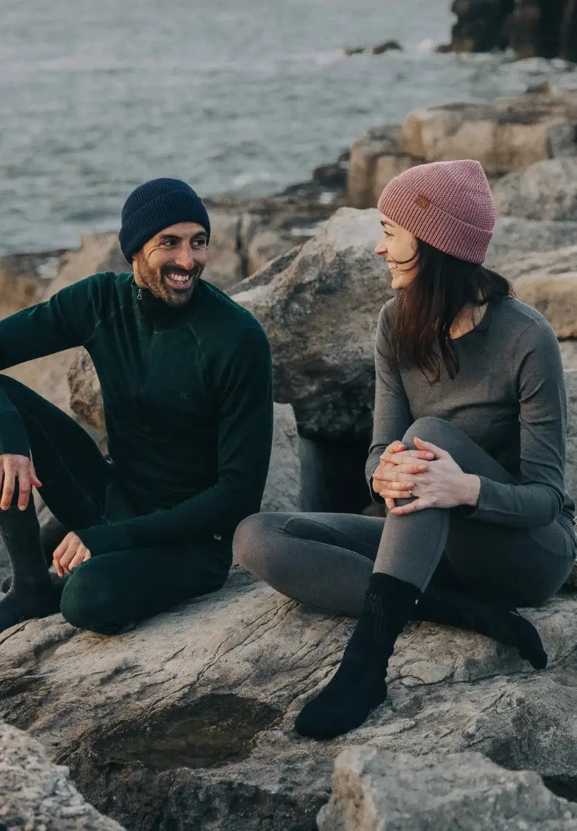 Two people in beanies sit on seaside rocks, smiling and wearing warm clothing in a natural setting.