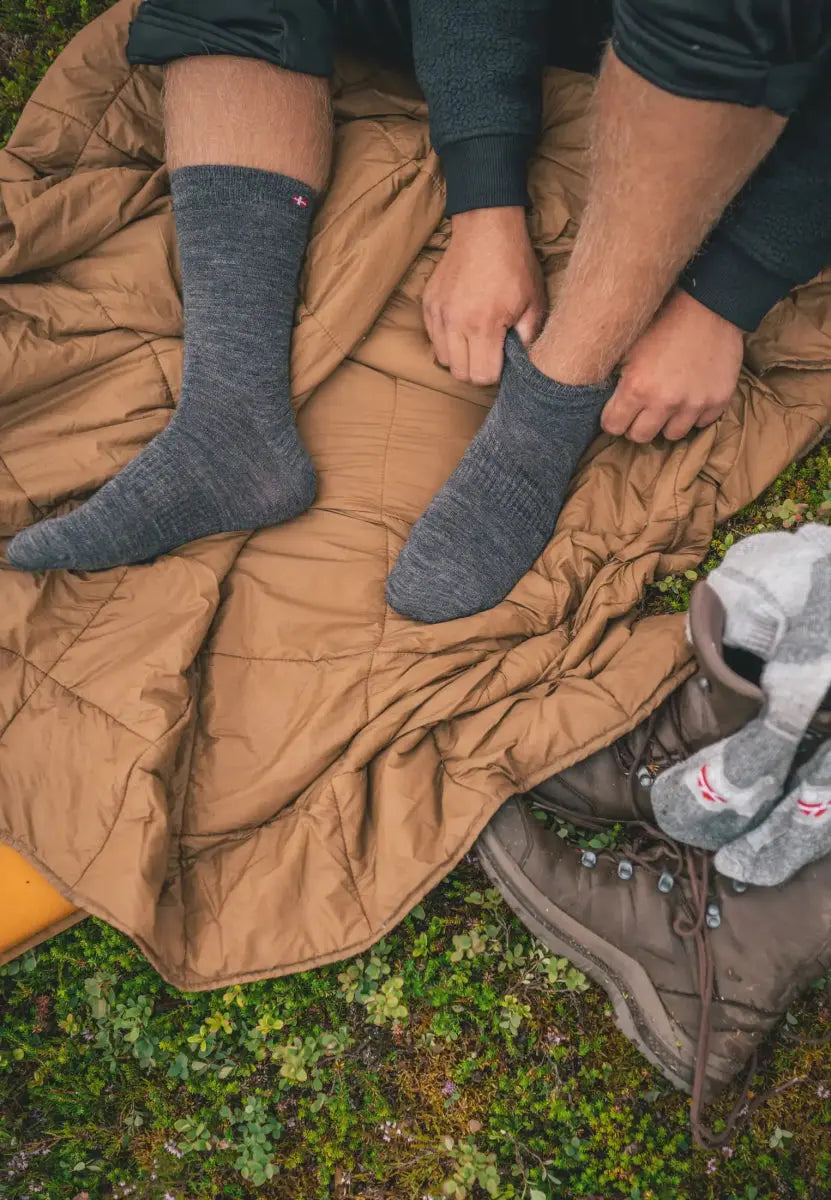 Person wearing gray socks on a tan blanket with hiking boots nearby on grass.