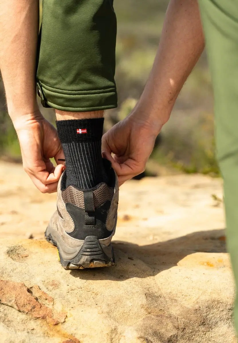Person adjusting black socks with emblem, wearing hiking shoes and green pants on rocky terrain.