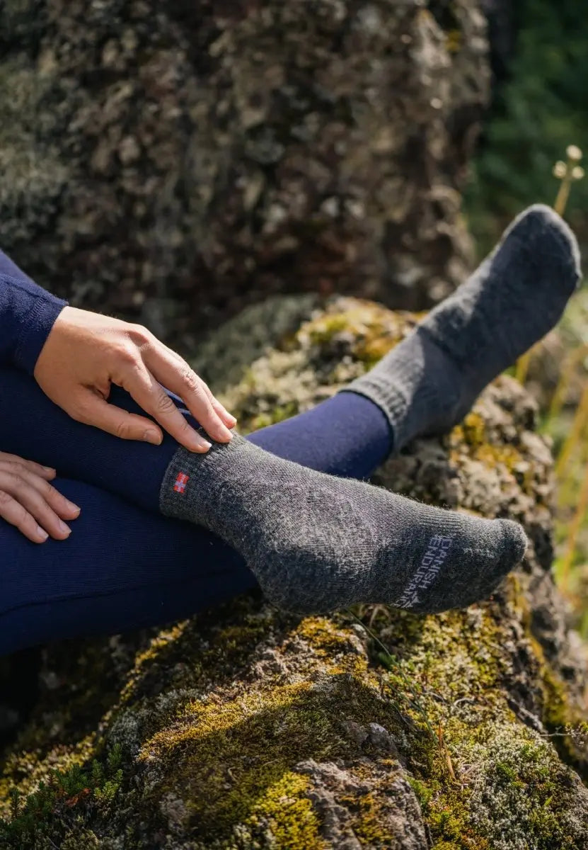 Person wearing gray hiking socks with emblem and text, sitting on moss-covered rock outdoors.