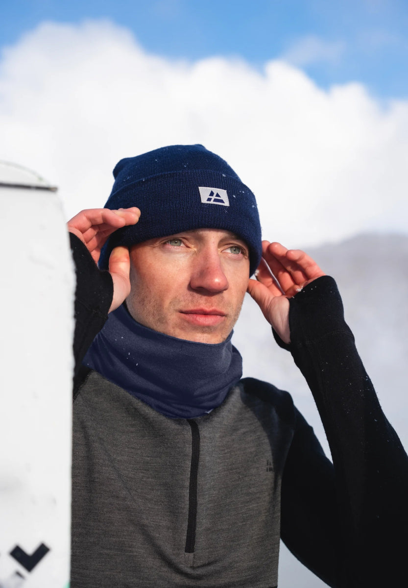 Person wears a blue beanie and neck gaiter against a snowy mountain backdrop.