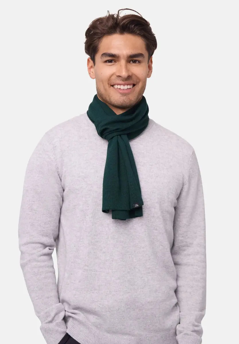 A person wears a green merino wool scarf with a light gray sweater, smiling warmly.