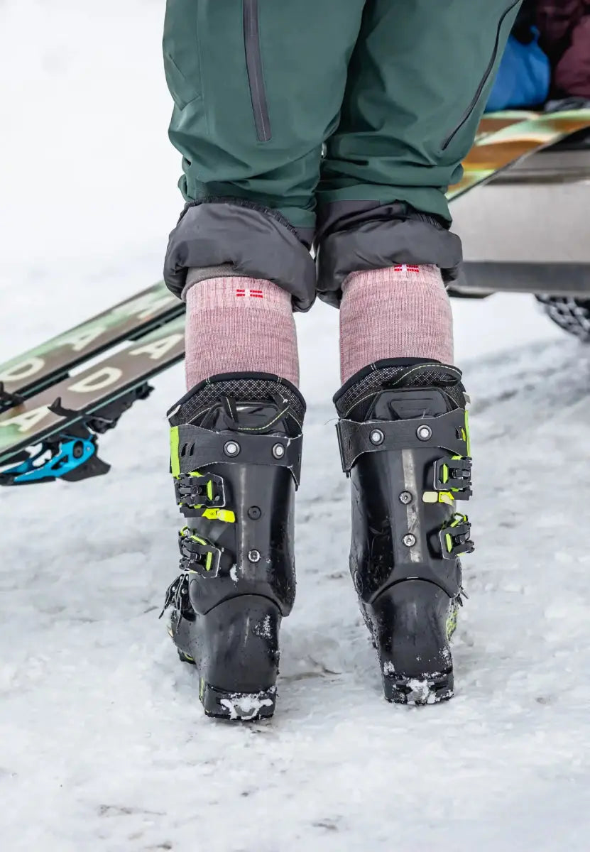 Person in ski boots wearing pink Merino wool socks, standing on snow with skis in the background.