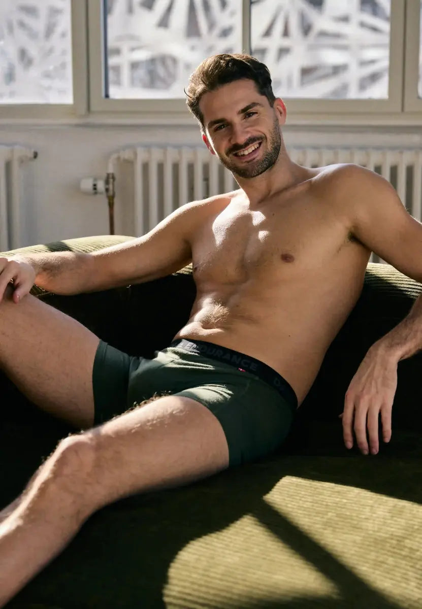 Man in dark green merino wool trunks, smiling while reclining on a couch, with sunlight streaming in.