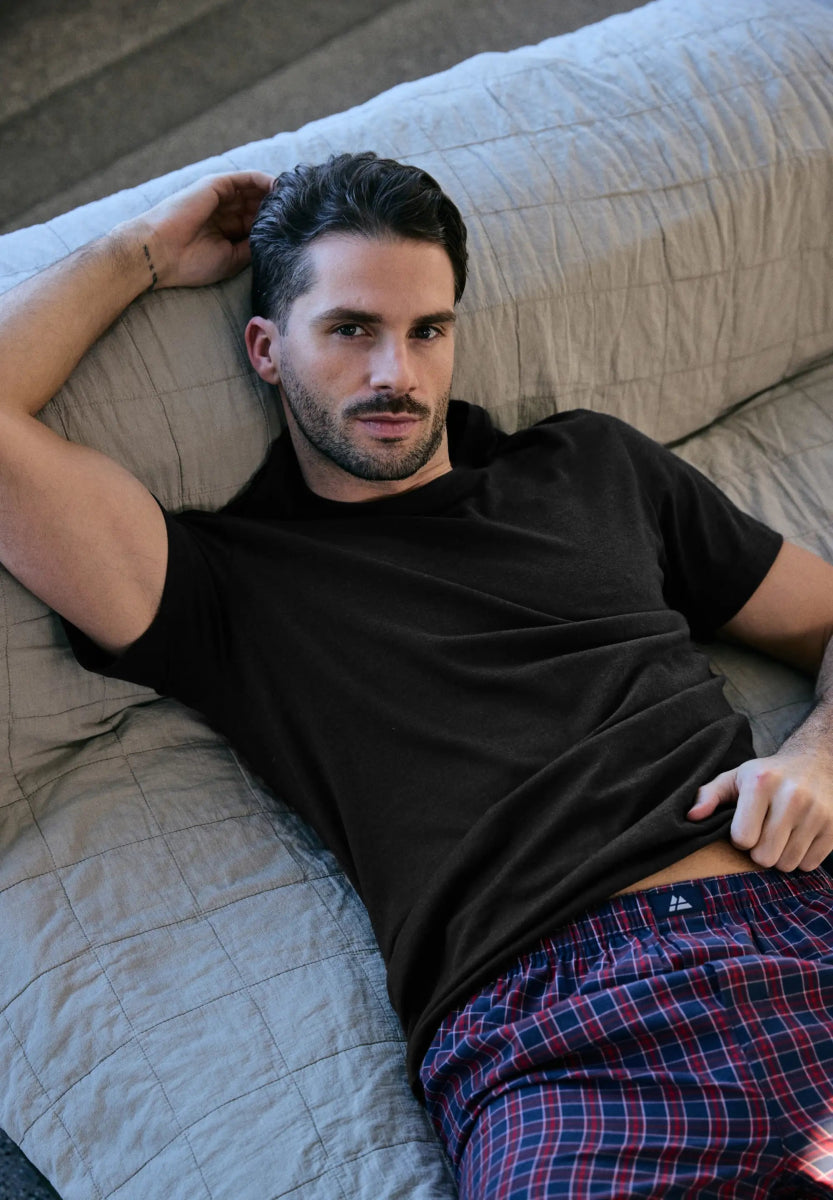 Man reclining on a couch, wearing a black crew neck t-shirt and plaid pajama pants.