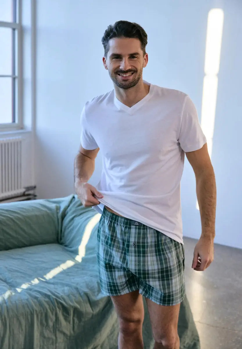 Man in white V-neck T-shirt and plaid shorts, smiling in a sunlit room with a green sofa.