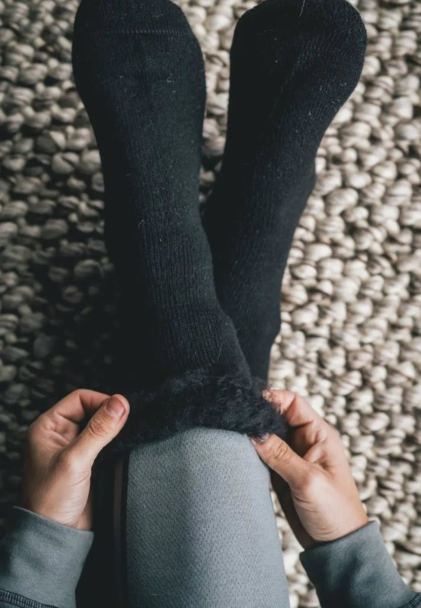 Black fuzzy socks being adjusted over gray pants on a textured rug.
