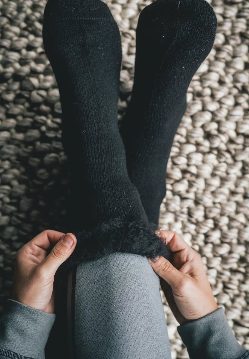 Hands adjusting fluffy black non-slip socks on legs, textured carpet background.